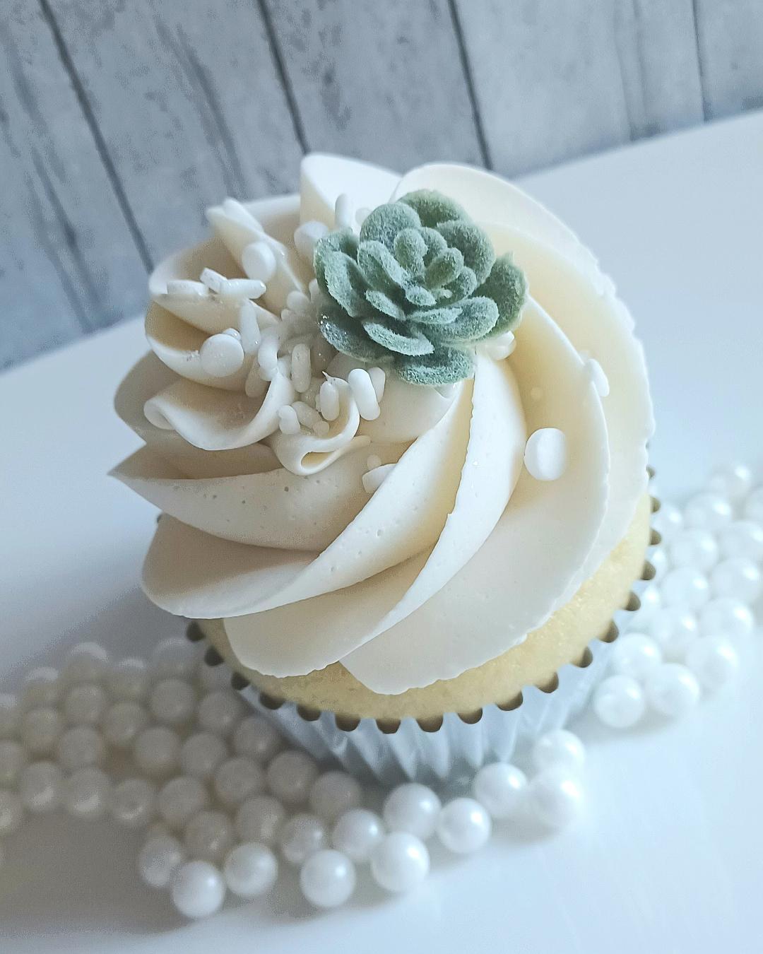 Cupcake with white frosting, a small succulent decoration, and white decorative balls around it, on a white surface.
