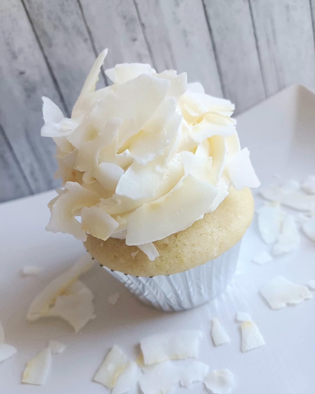 Cupcake with white frosting and white chocolate shavings on top, on a white surface with scattered chocolate pieces, background of wooden panels.