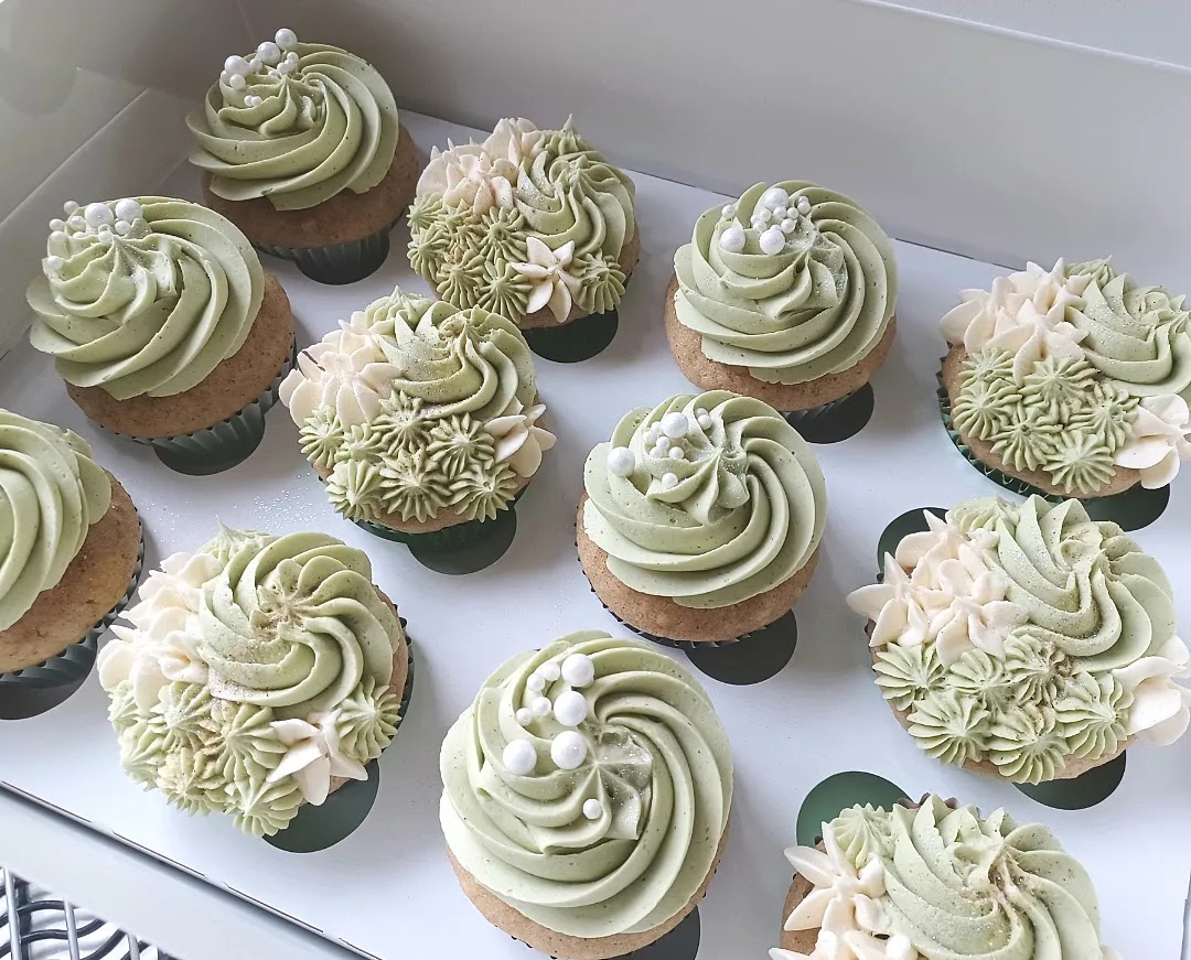 Box of decorated cupcakes with white and green frosting, floral and swirl designs, some with pearl-like accents.