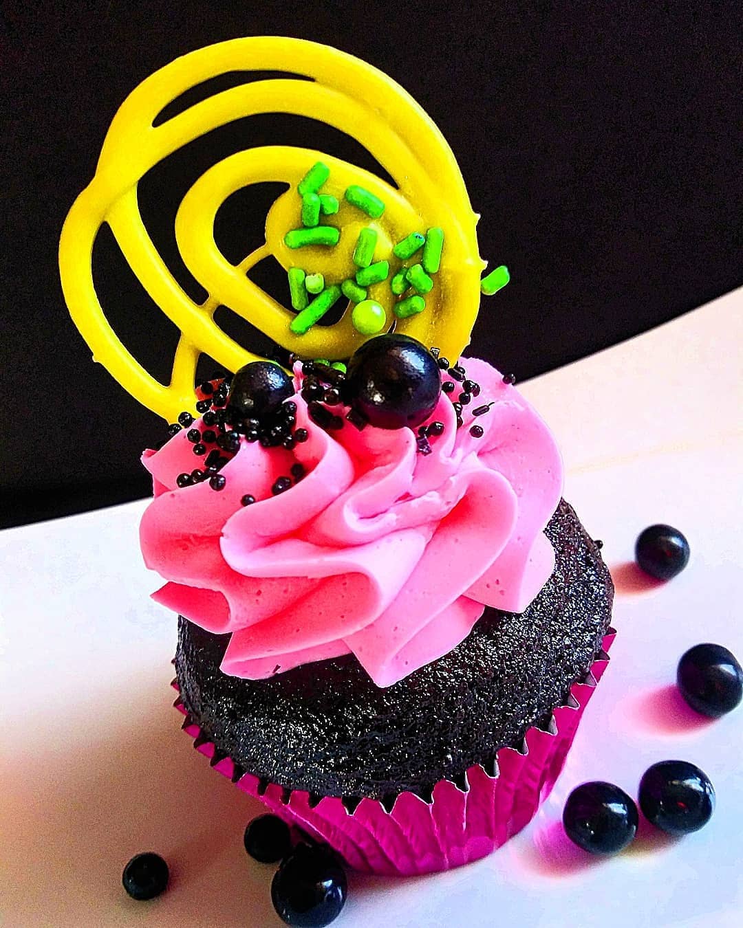 Cupcake with pink frosting, black cake, and black berries, topped with yellow spiral decoration and green sprinkles, on a white surface.