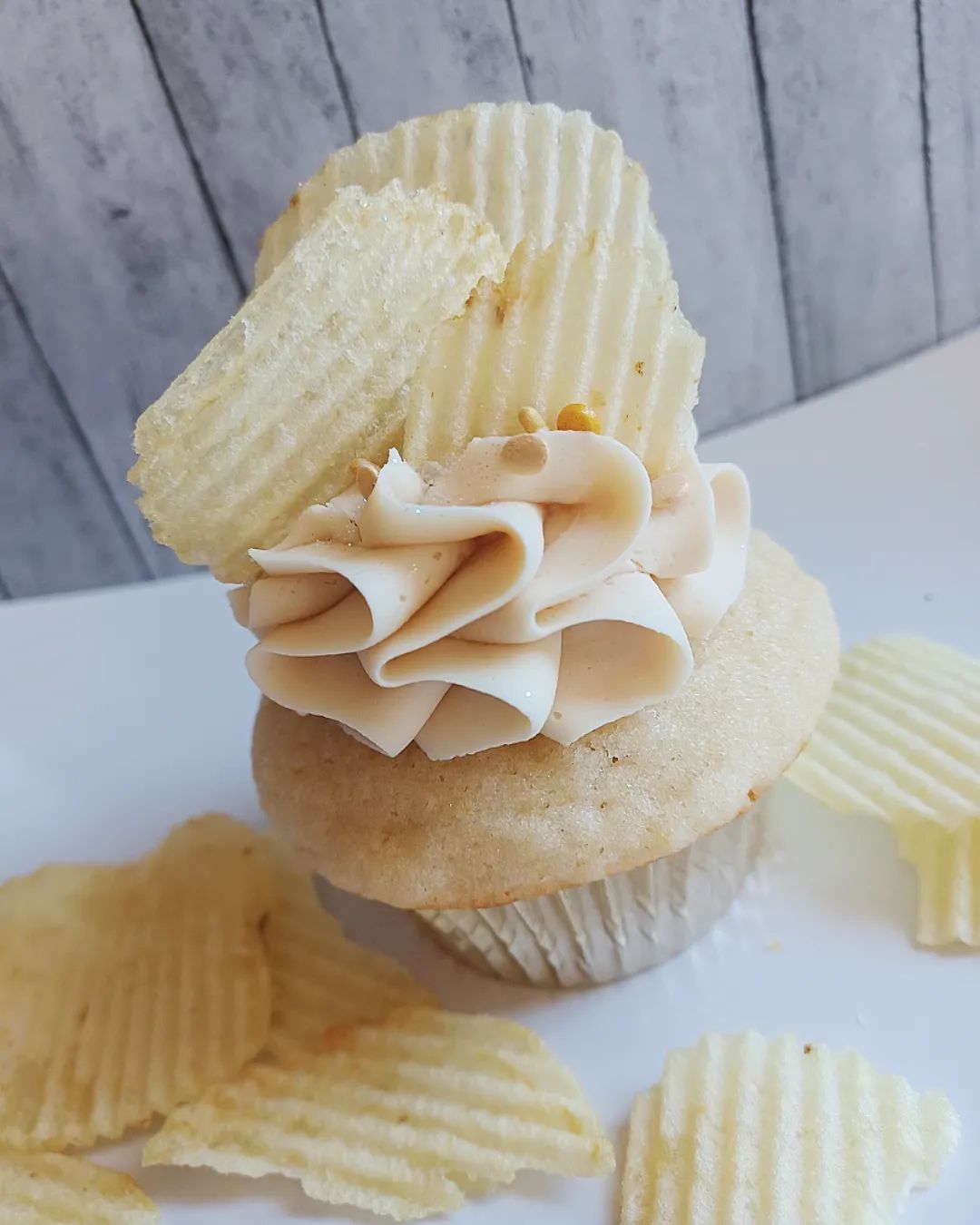 Cupcake with cream, potato chips, and a cookie on top, with additional chips around it, against a wooden background.