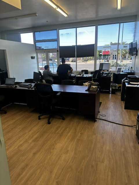 Office with large windows, two people sitting at desks, multiple chairs, and a wooden floor.