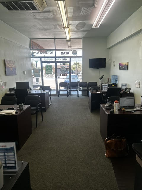 Office space with desks, chairs, a wall-mounted TV, and large front windows showing parked cars outside.