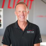 Smiling man in black polo shirt standing indoors with a blurred background and red text. Word 'FLIGHT' visible.