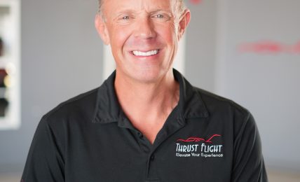 Smiling man in black polo shirt standing indoors with a blurred background and red text. Word 'FLIGHT' visible.