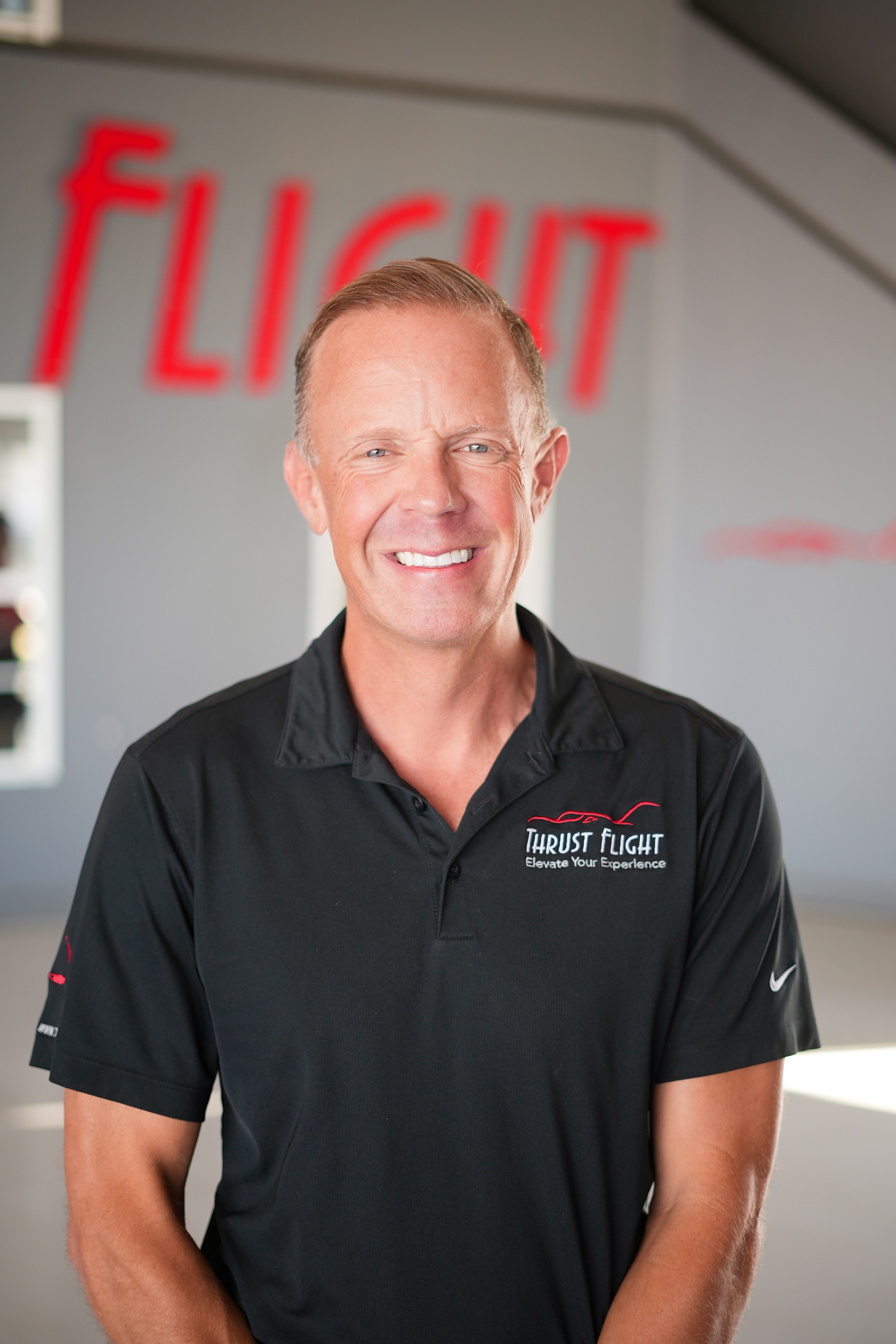 Smiling man in black polo shirt standing indoors with a blurred background and red text. Word 'FLIGHT' visible.