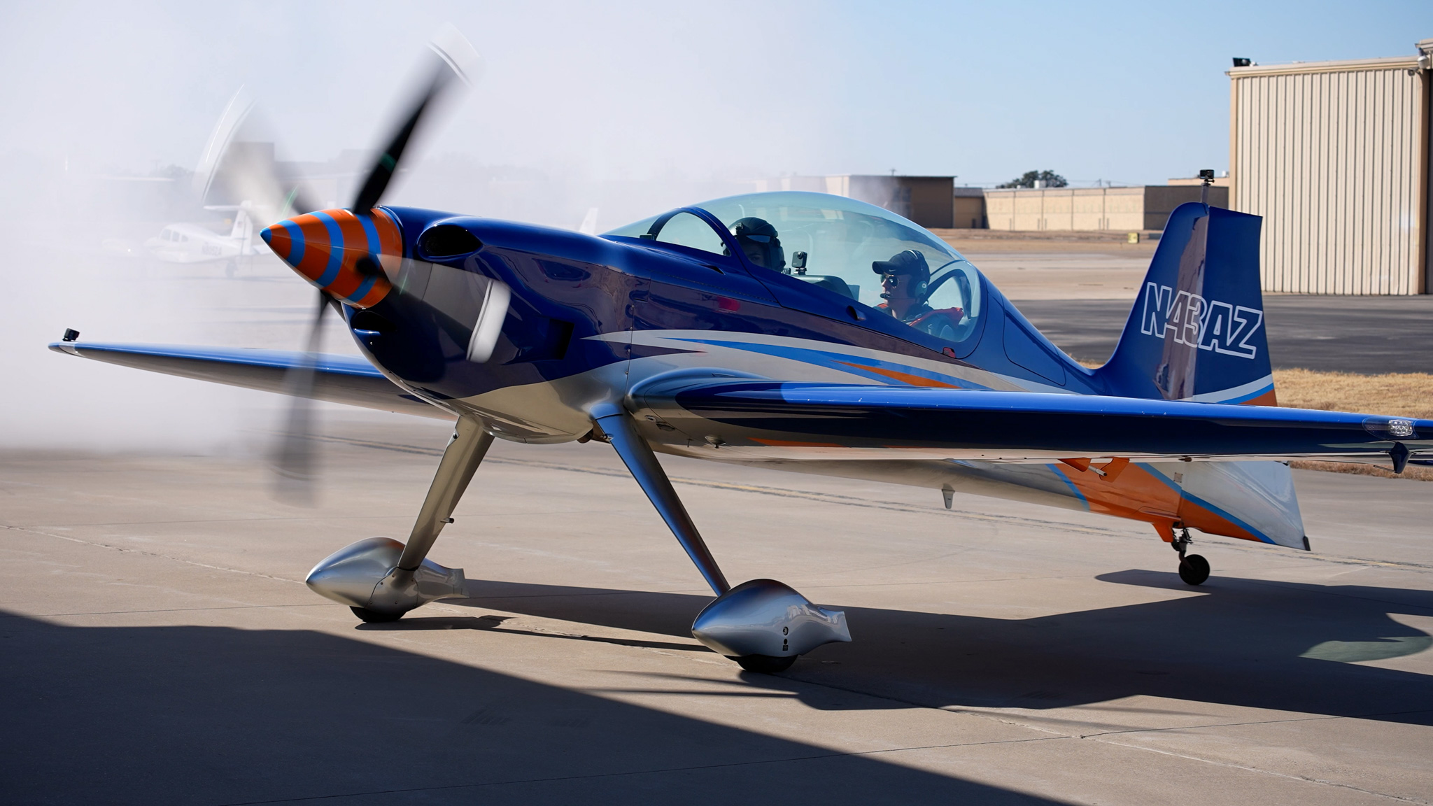 Small blue and orange propeller aircraft on tarmac with hangar in background.