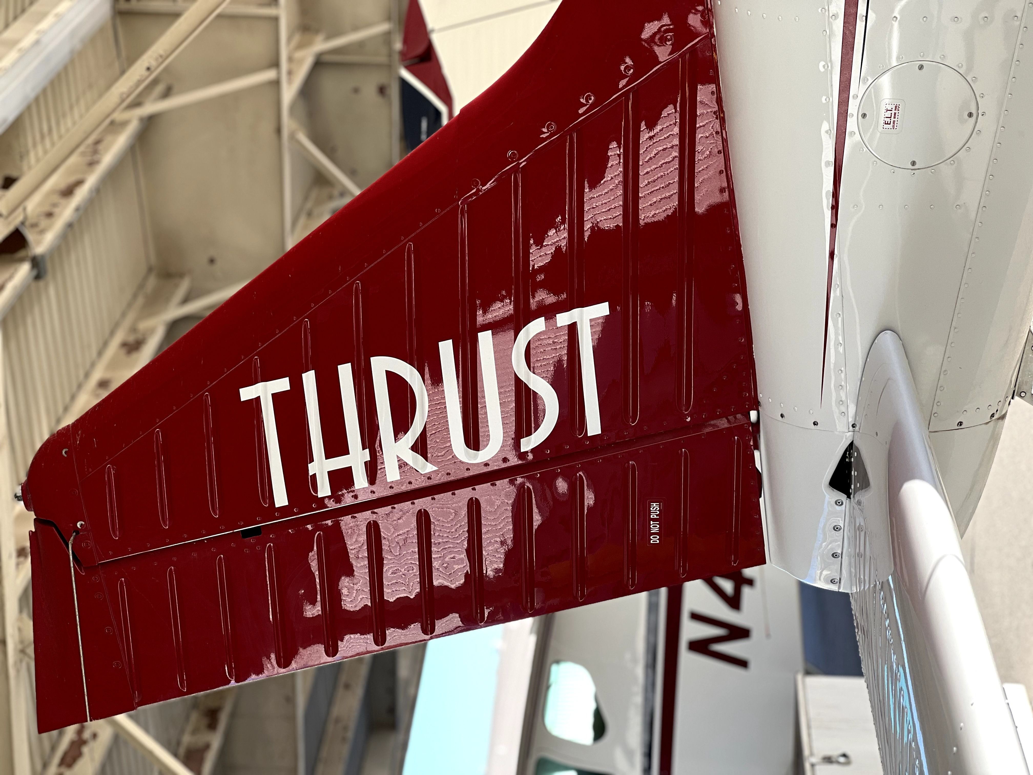 Red airplane tail with white text 'THRUST' inside a hangar, with part of another aircraft visible in the background.