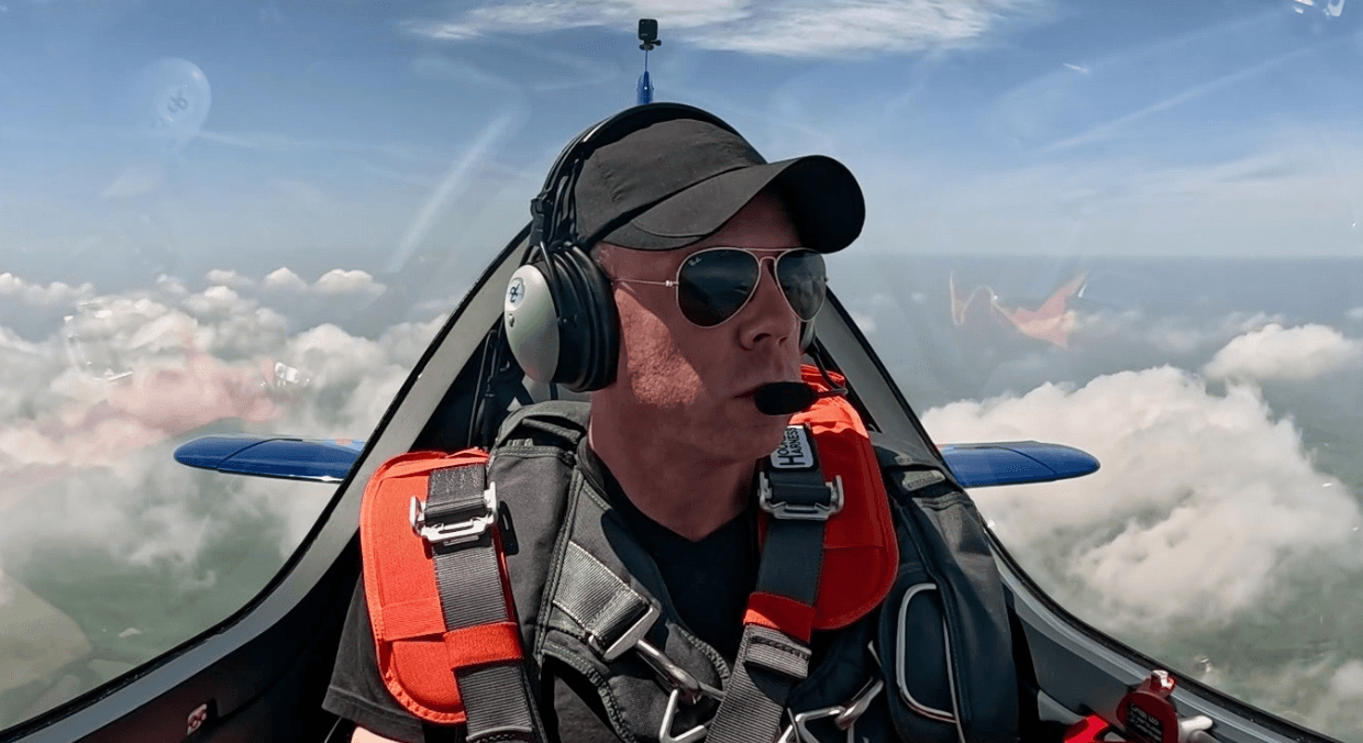 Person wearing sunglasses, headset, and harness in an aircraft cockpit flying above clouds.