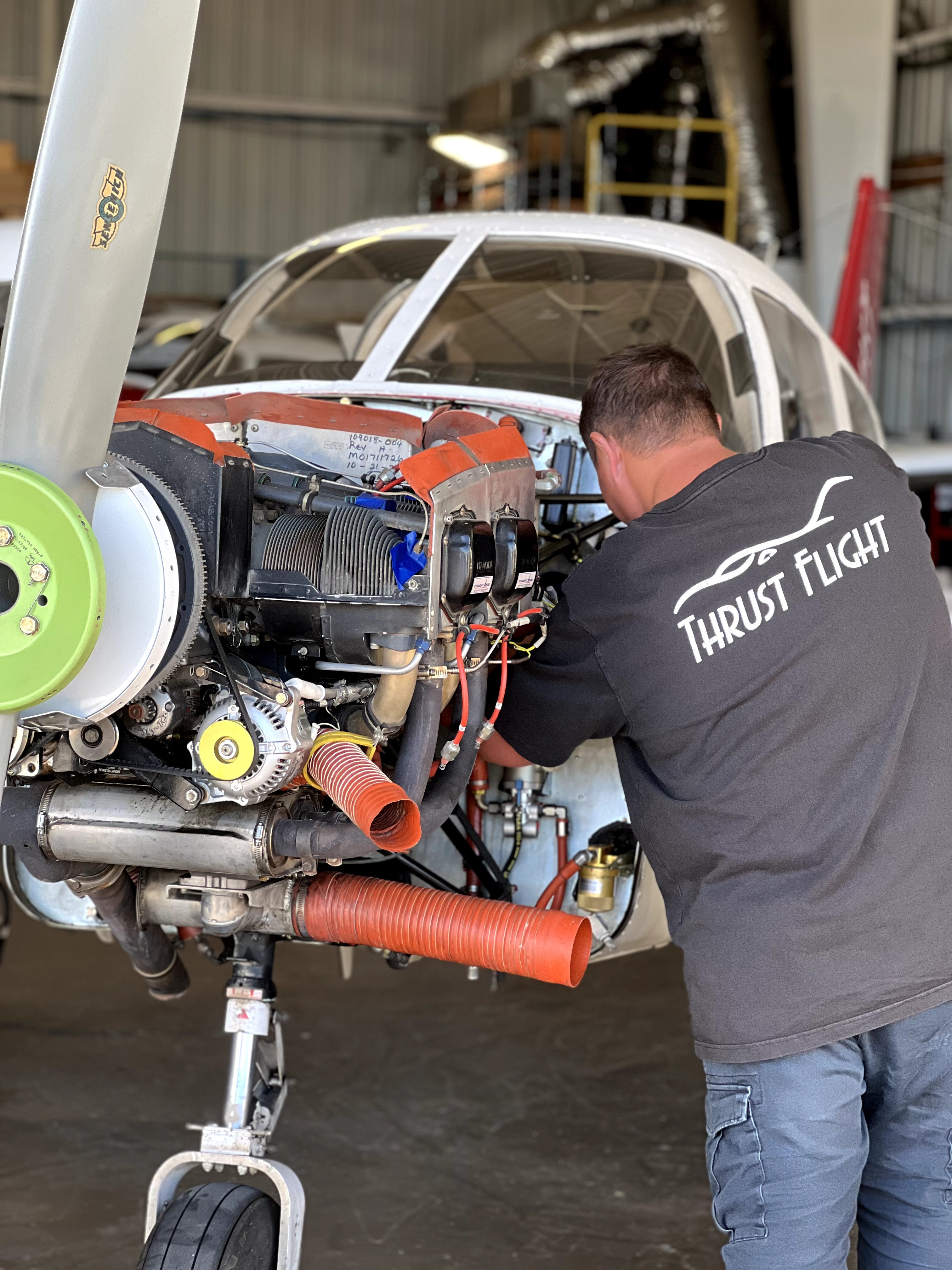 Person working on aircraft engine inside hangar, wearing a black shirt with 'Thrust Flight' logo, with aircraft cockpit visible.