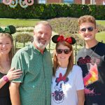 Four people smiling outdoors, two women and two men, with a green hedge and decorative elements in the background.