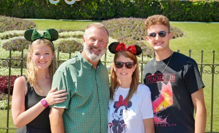 Four people smiling outdoors, two women and two men, with a green hedge and decorative elements in the background.