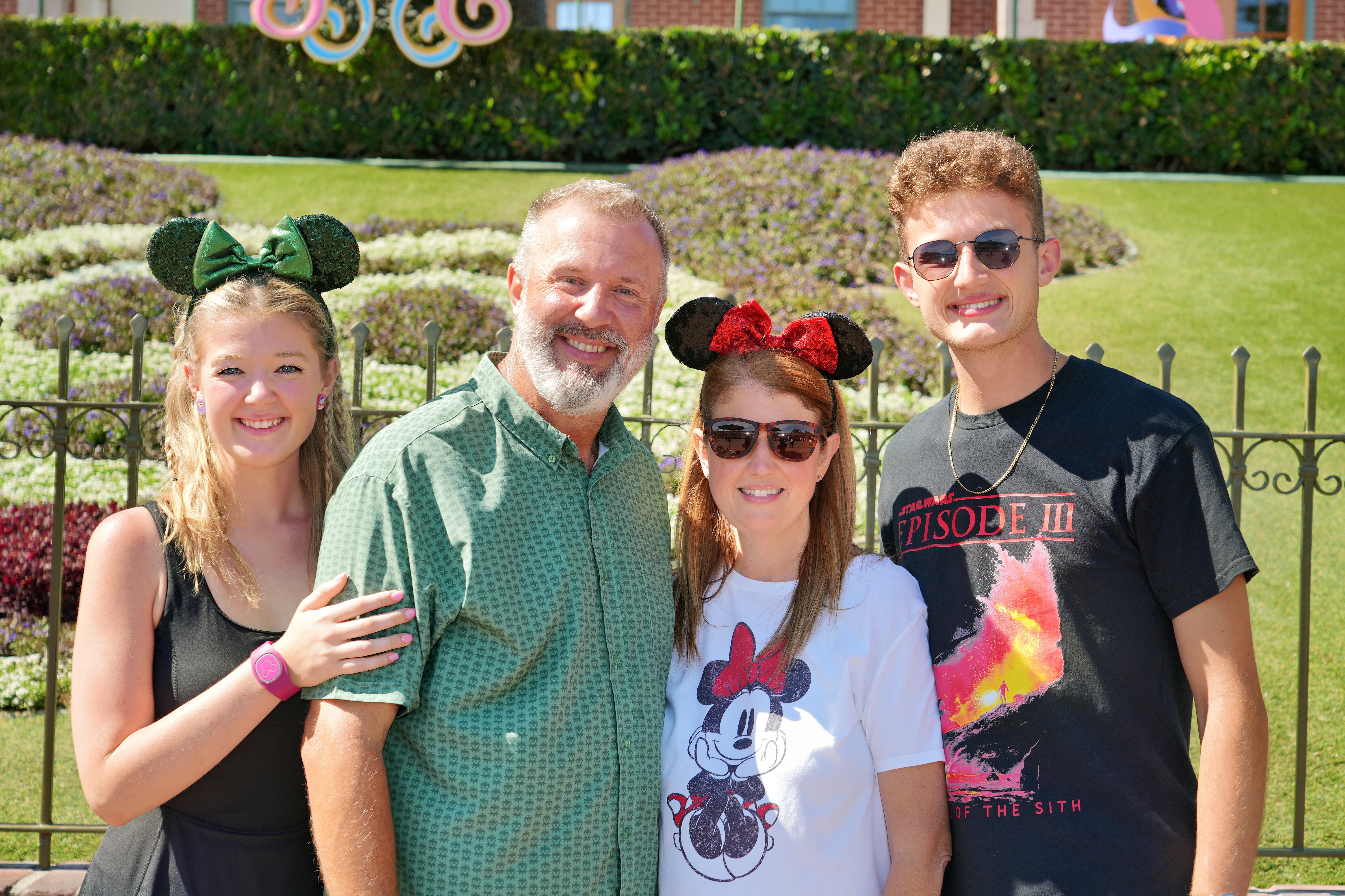 Four people smiling outdoors, two women and two men, with a green hedge and decorative elements in the background.