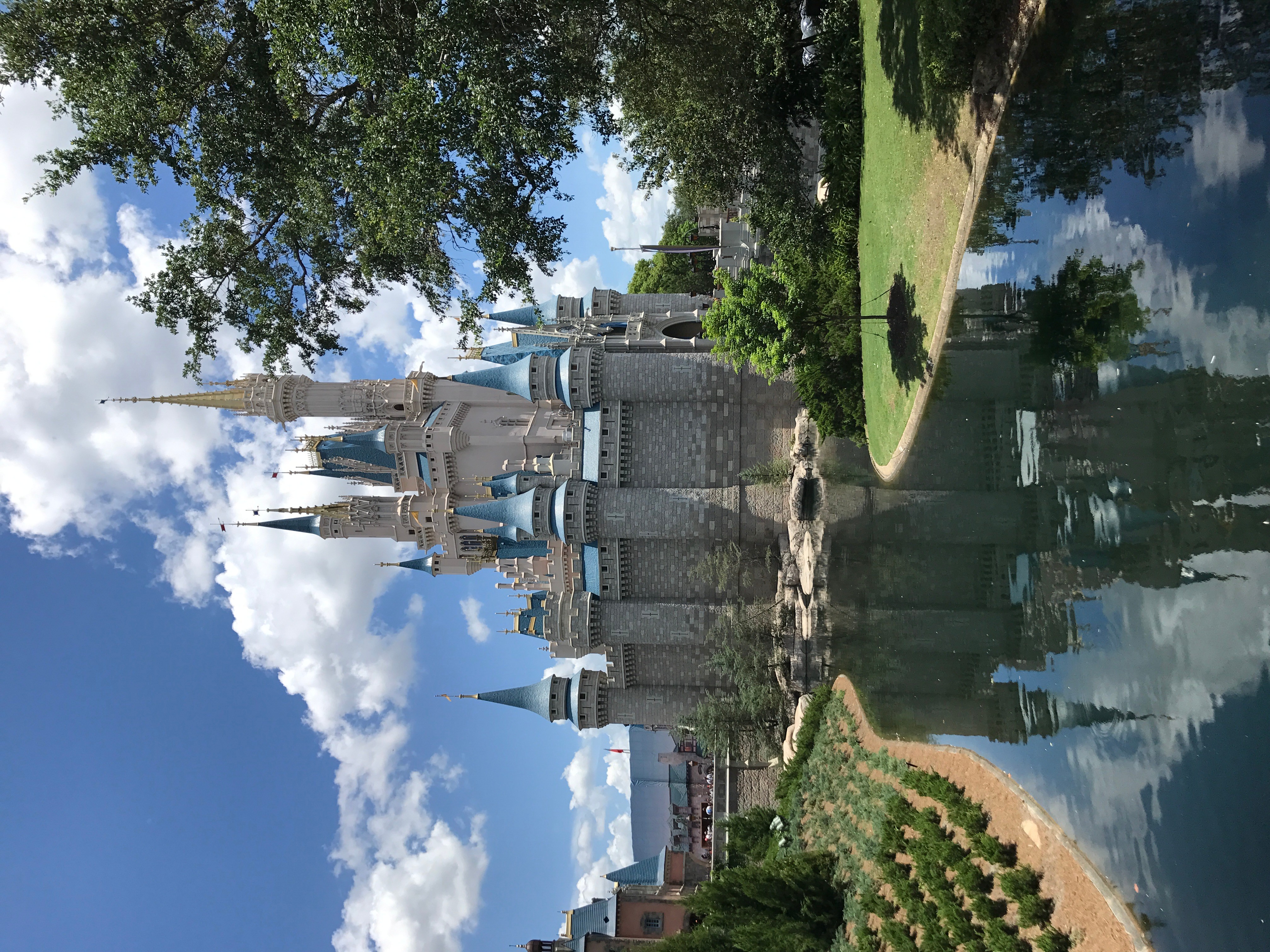 Castle with blue turrets reflected in a pond, surrounded by trees and a grassy area, under a partly cloudy sky.