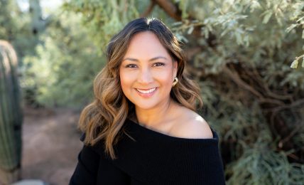 Woman with wavy hair smiling outdoors, wearing a black off-shoulder top, surrounded by greenery.