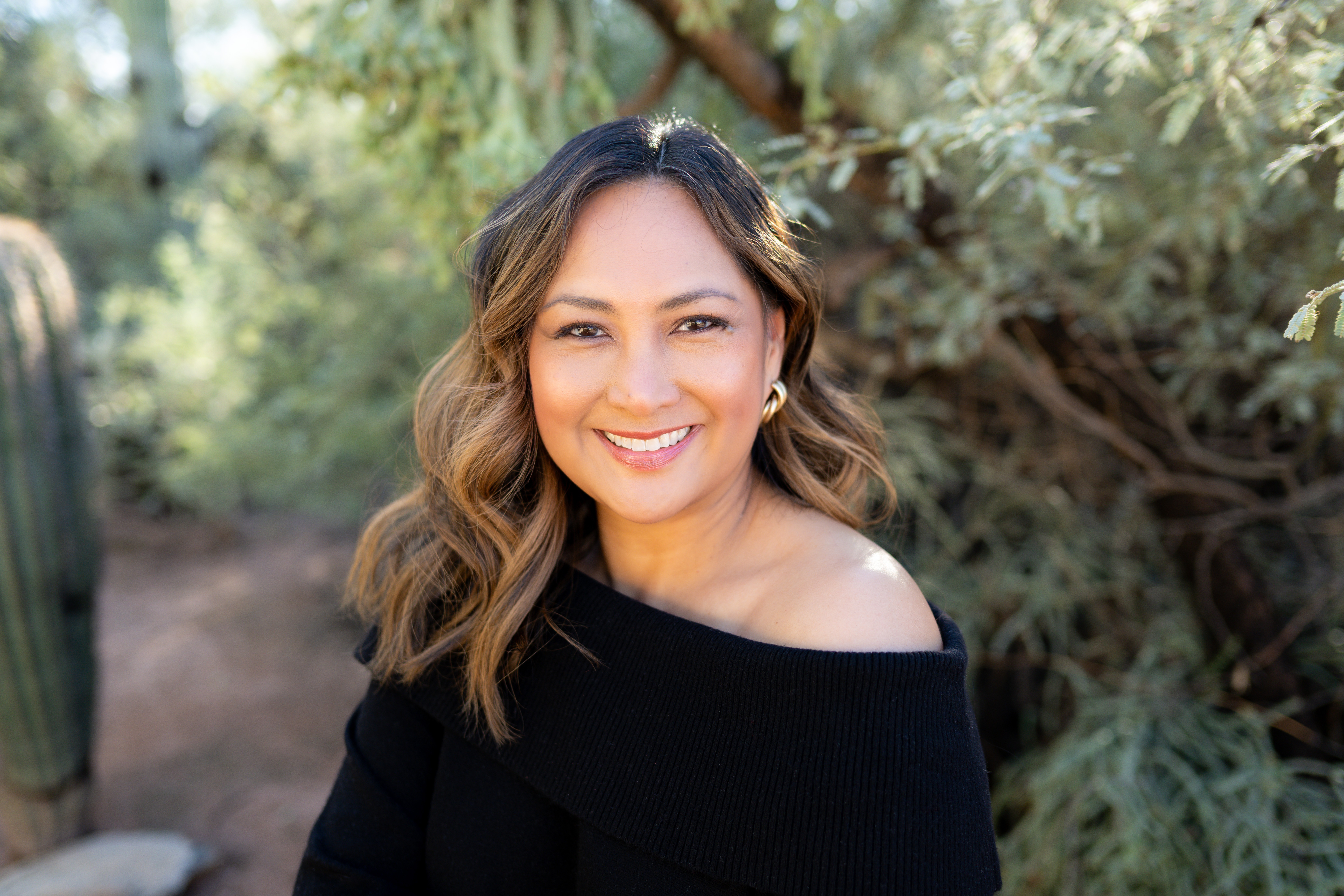 Woman with wavy hair smiling outdoors, wearing a black off-shoulder top, surrounded by greenery.