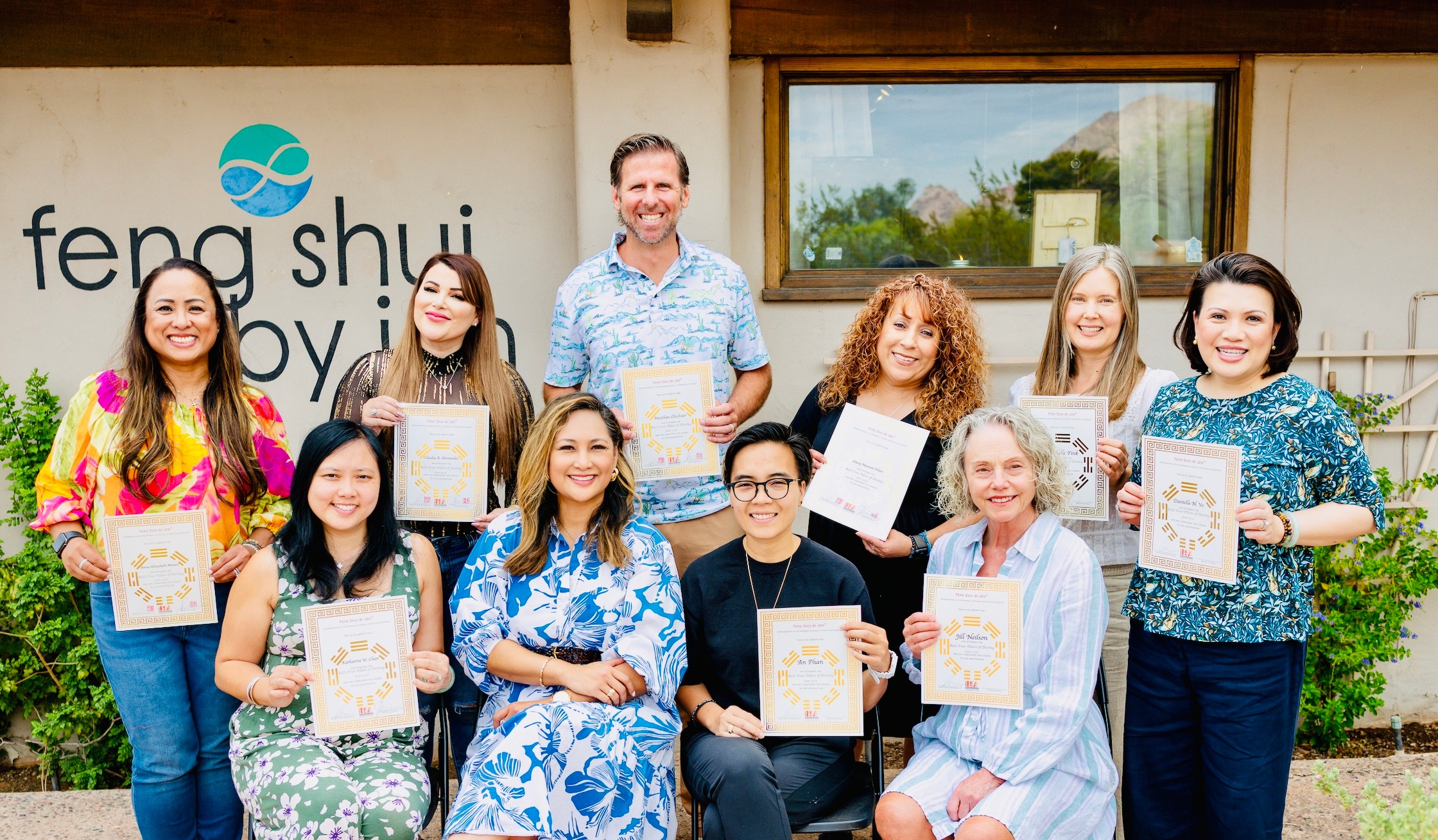 Group of ten people holding certificates outside a building with a window and greenery, smiling for the photo.