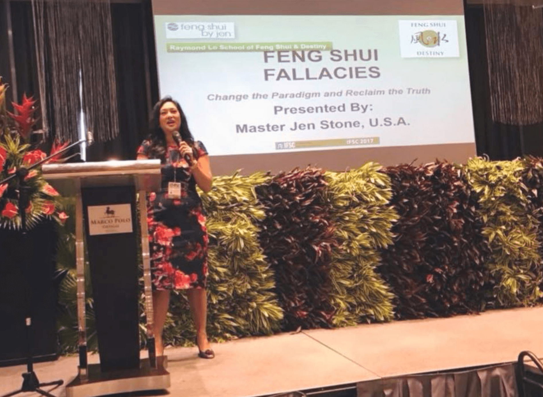 Woman standing on stage in front of a presentation screen with plants and flowers in front, giving a talk.