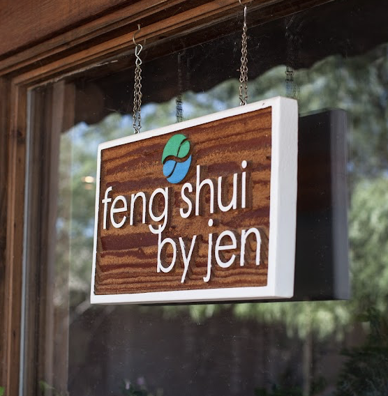 Wooden sign hanging outside with text 'feng shui by jen' and a logo above, reflected in a window.