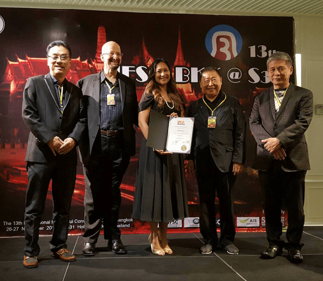 Five people standing on stage holding a certificate, with a large screen behind them displaying event information.