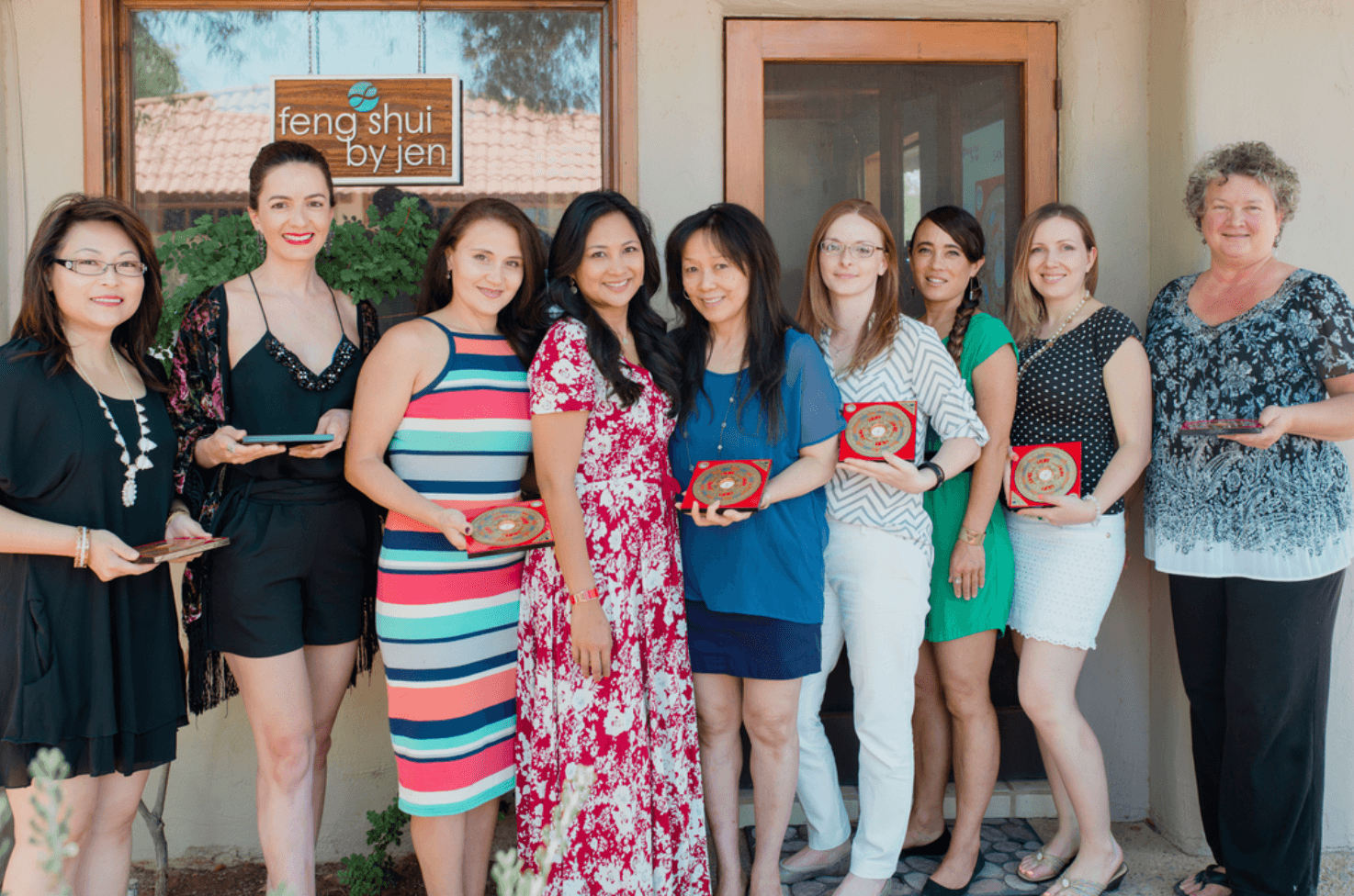 Group of nine women standing outside a building, some holding awards, smiling, with a sign in the window behind them.