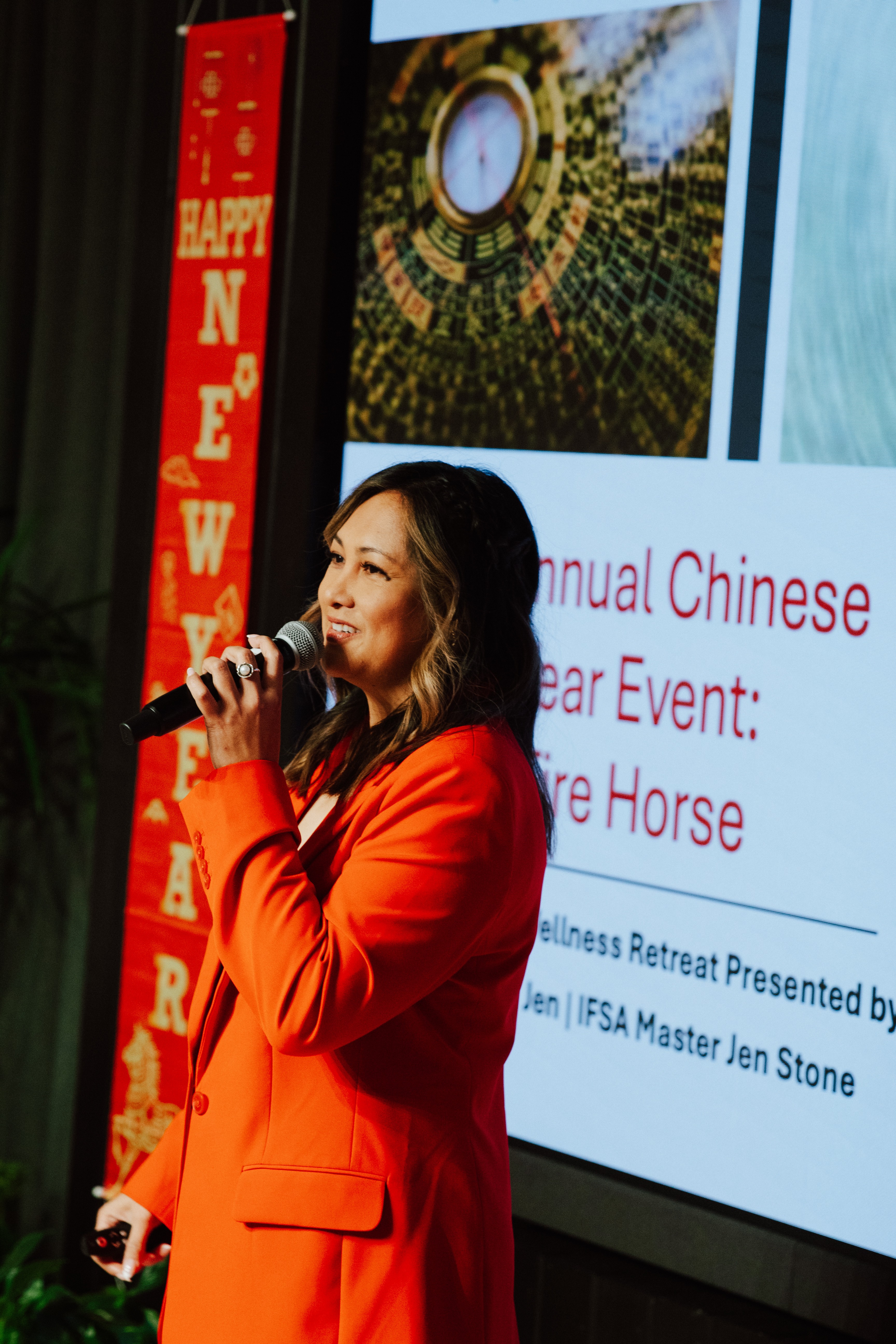 Woman in red blazer speaking into microphone at event, with presentation screen behind her displaying text and images.