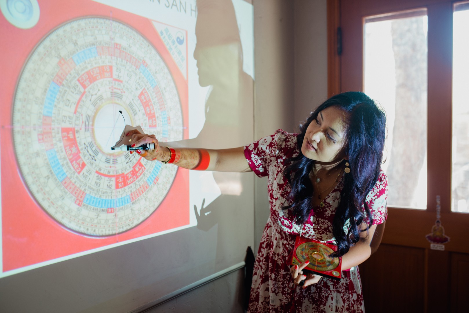 Woman pointing at a colorful circular diagram projected on a wall, holding a marker and a notebook, indoors with a window.