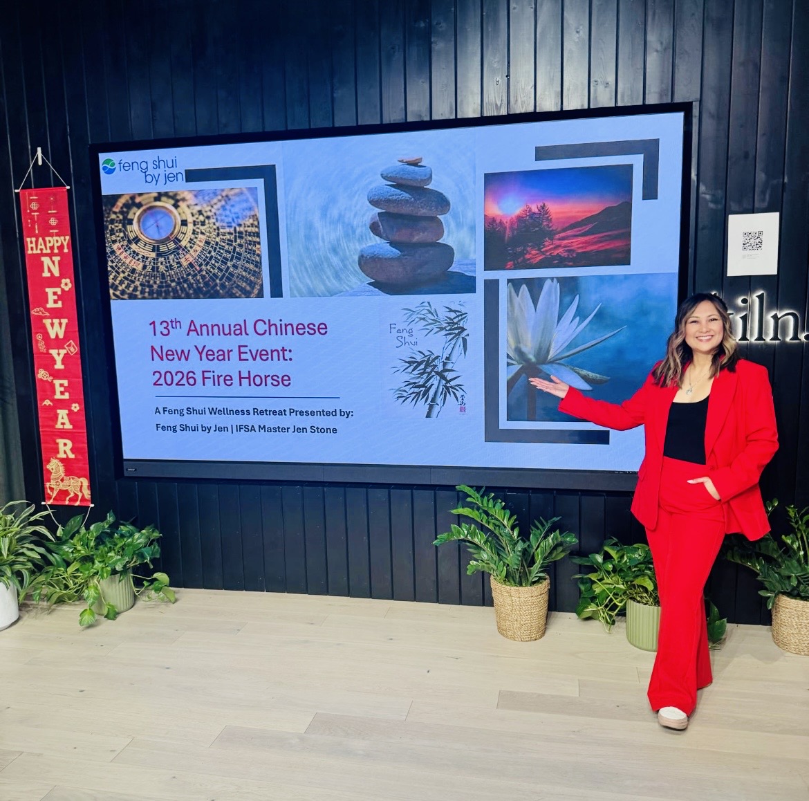 Woman in red suit standing next to a large screen displaying event information, with plants and a New Year's banner nearby.