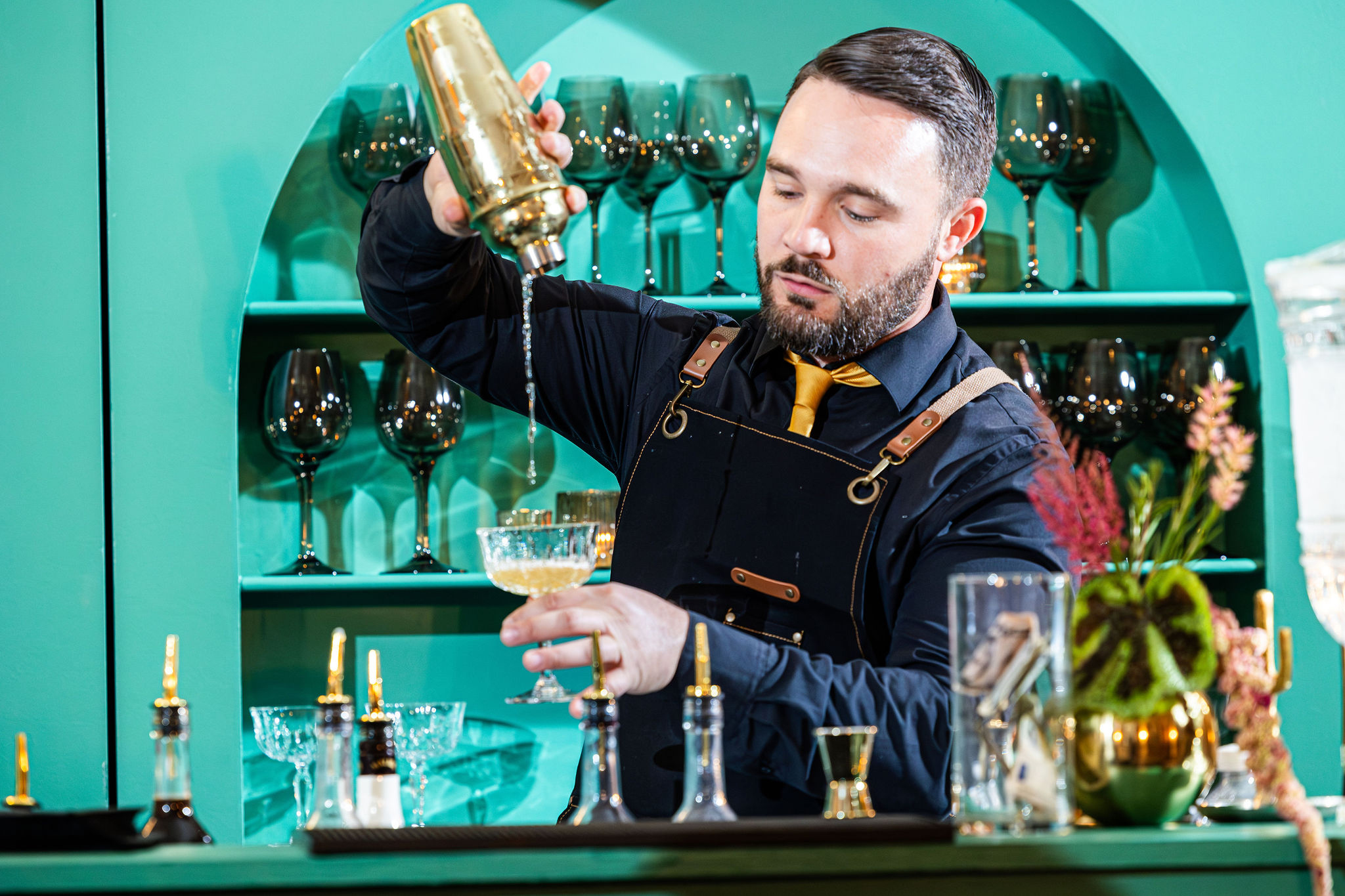 Bartender pouring a drink into a glass, with shelves of wine glasses behind him, in a colorful bar setting.