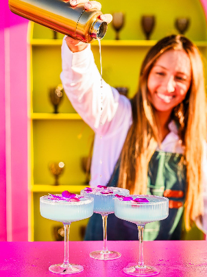 Woman pouring liquid into three glasses with pink and purple decorations, smiling, colorful background with shelves and trophies.
