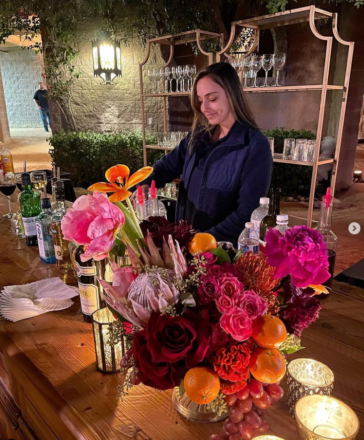 Woman preparing drinks at a bar with a large floral arrangement and bottles on a wooden table, outdoor setting at night.