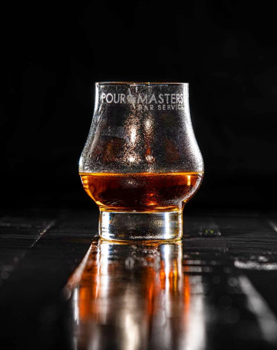 Glass with dark amber liquid on a reflective surface against a black background, condensation on the glass, and text partially visible.