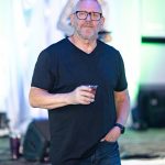 Man with glasses and a beard holding a drink, wearing a black t-shirt, at an indoor event with colorful lighting.