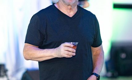 Man with glasses and a beard holding a drink, wearing a black t-shirt, at an indoor event with colorful lighting.