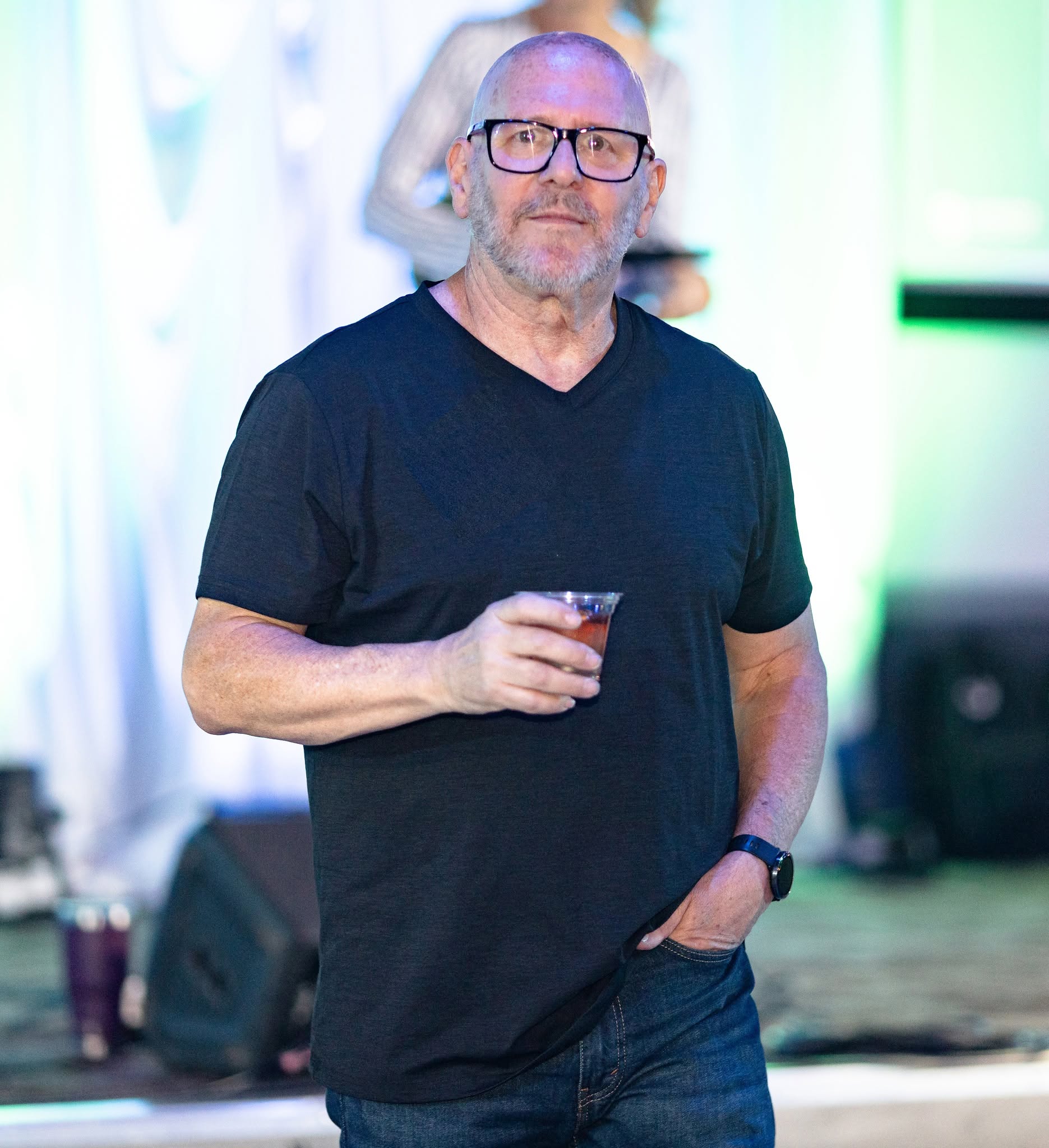 Man with glasses and a beard holding a drink, wearing a black t-shirt, at an indoor event with colorful lighting.