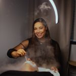 Smiling woman with dark hair in a black top, sitting in a room with a circular light above, steam rising from a device.