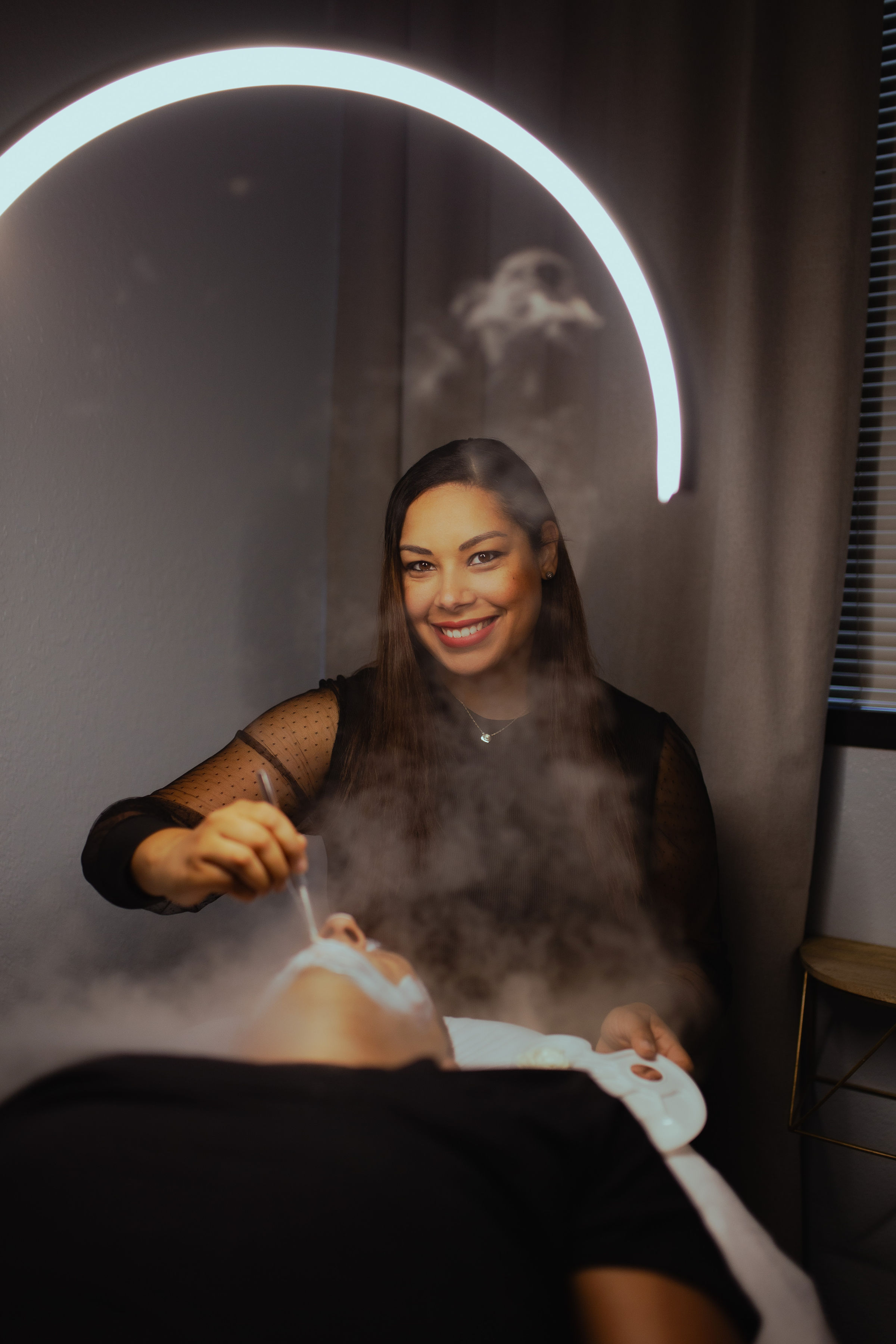 Smiling woman with dark hair in a black top, sitting in a room with a circular light above, steam rising from a device.