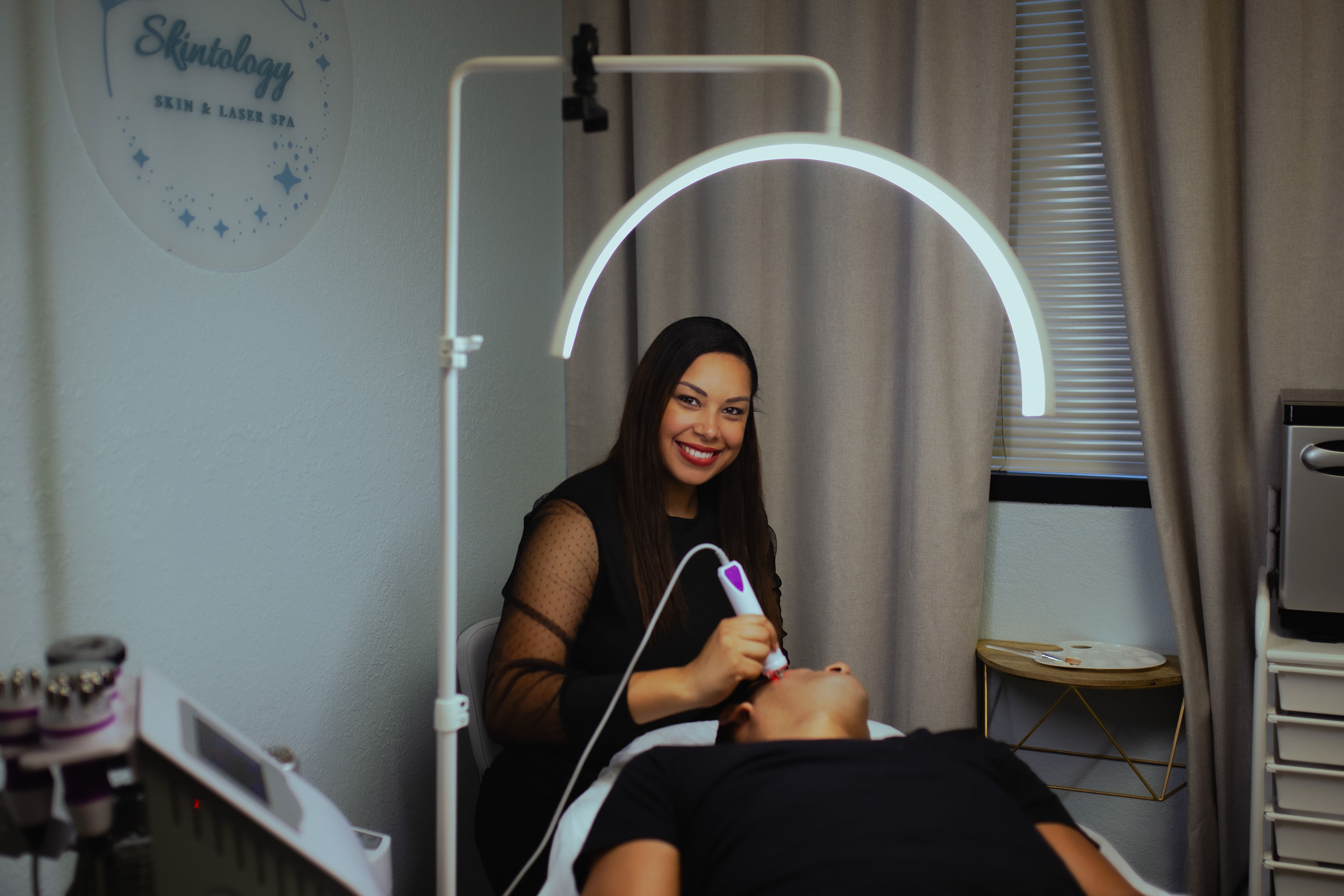 Smiling woman in black dress holding a device, seated in a medical or dental setting with equipment and curtains nearby.