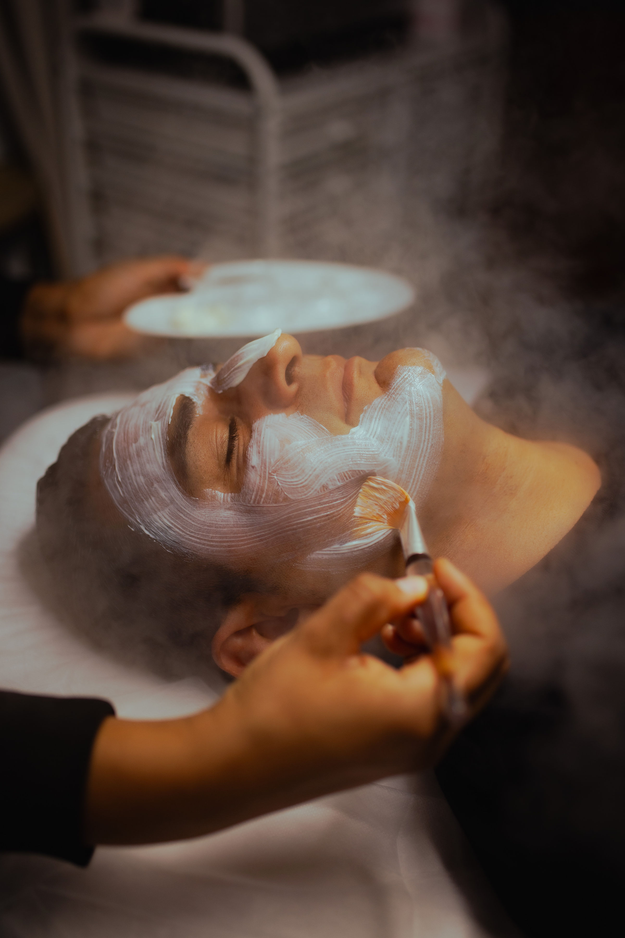 Person receiving facial treatment with steam and mask, lying with eyes closed, in a spa setting.