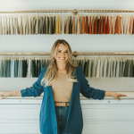 Woman standing in fabric store with colorful fabric swatches on wall behind her, smiling, arms outstretched, in a well-lit shop.
