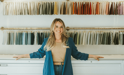 Woman standing in fabric store with colorful fabric swatches on wall behind her, smiling, arms outstretched, in a well-lit shop.