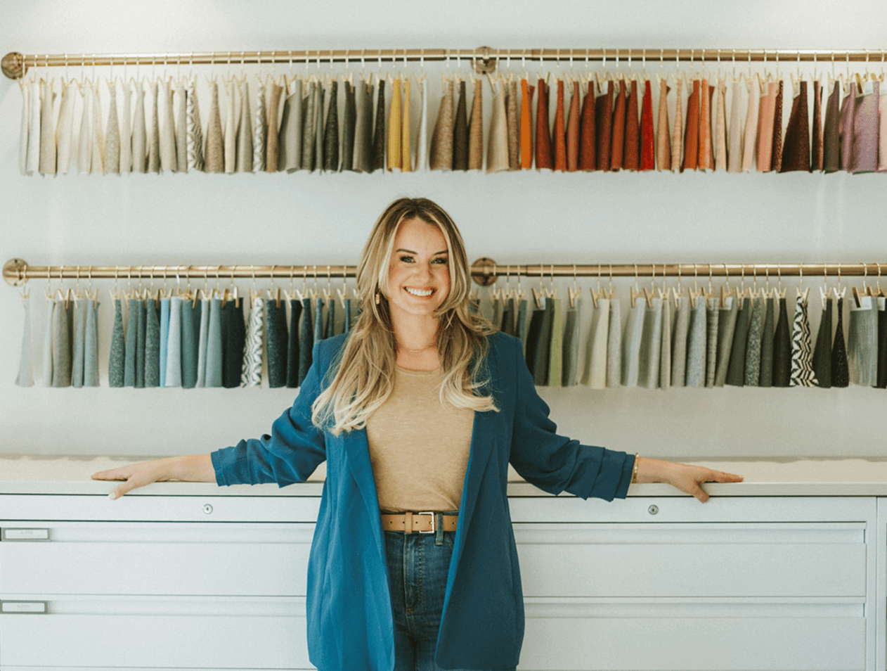 Woman standing in fabric store with colorful fabric swatches on wall behind her, smiling, arms outstretched, in a well-lit shop.
