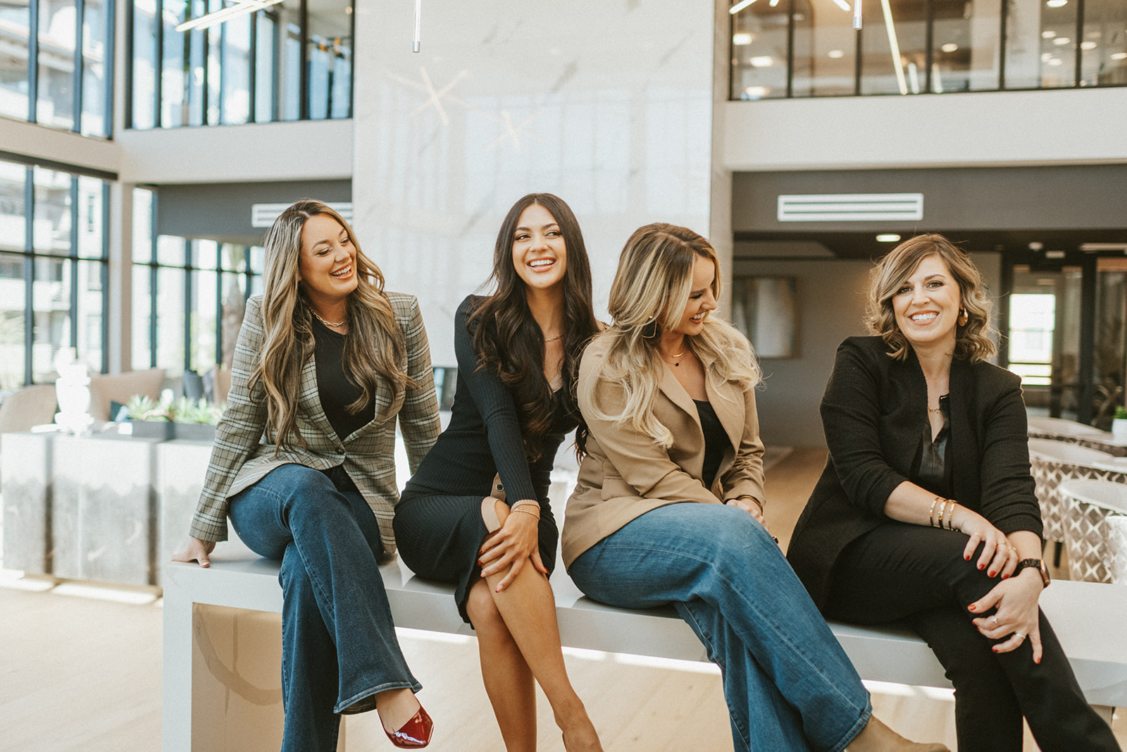 Four women sitting on a white bench in a modern indoor space, smiling and talking, with large windows behind them.