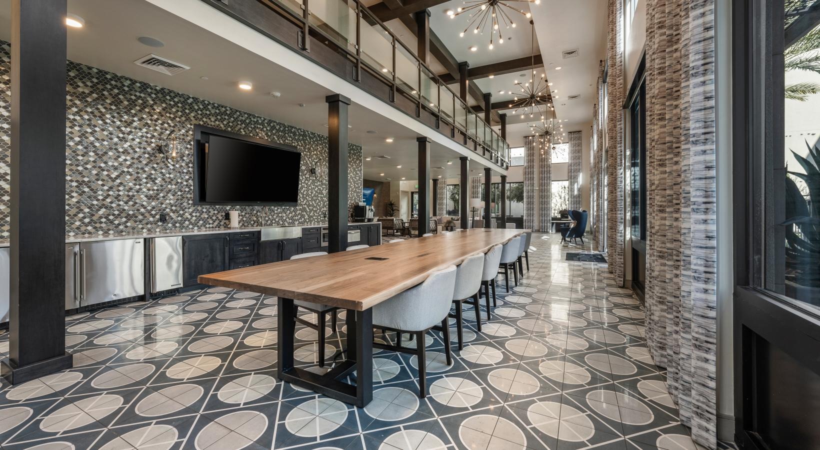 Modern dining area with a long wooden table, white chairs, patterned floor, large windows, and a wall-mounted TV.