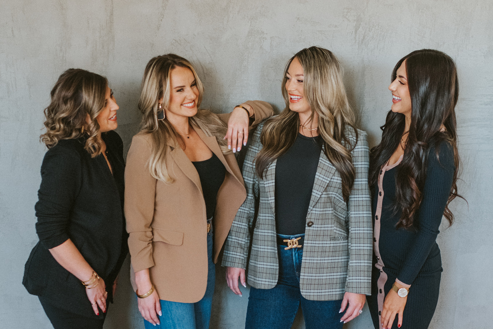 Four women standing together, smiling and talking, against a plain gray wall, dressed in casual and business attire.